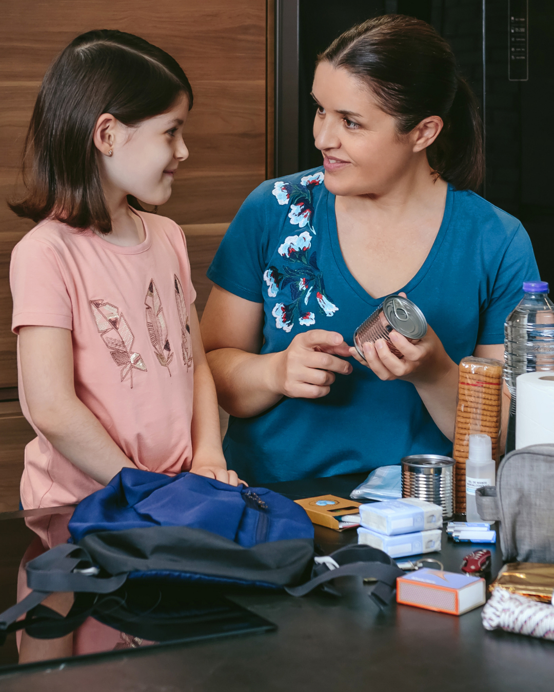 a woman holds a can of food, around other preparedness items, with a young girl